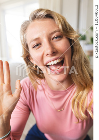 Happy blonde caucasian woman having video call and waving hand in sunny living room Happy blonde caucasian woman having video call and waving hand in sunny living room 110040012