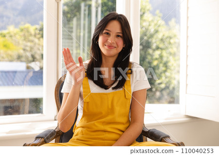 Happy biracial teenage girl having video call sitting in sunny room waving Happy biracial teenage girl having video call sitting in sunny room waving 110040253