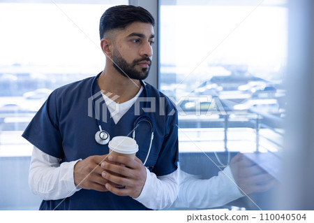 Biracial male doctor using smartphone and holding cup of coffee in hospital office 110040504