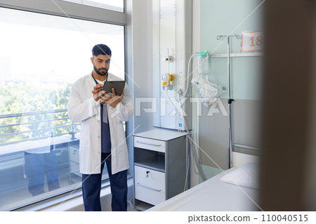 Biracial male doctor using tablet in sunny hospital room 110040515