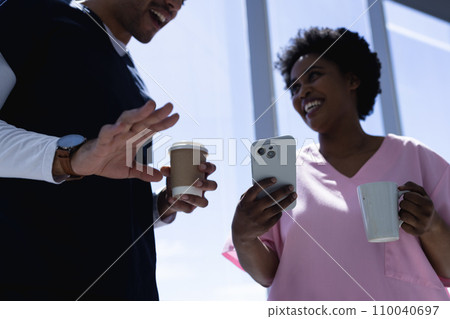 Happy diverse male and female doctors talking, drinking coffee and using smartphone in hospital 110040697