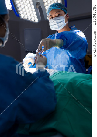 Diverse female doctor with face mask doing surgery in group of doctors in hospital operating room Diverse female doctor with face mask doing surgery in group of doctors in hospital operating room 110040786