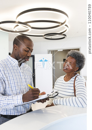 African american male doctor taking notes and talking with senior woman in hospital reception 110040789