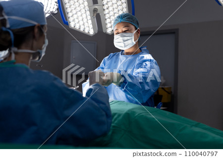 Diverse female doctor with face mask doing surgery in group of doctors in hospital operating room Diverse female doctor with face mask doing surgery in group of doctors in hospital operating room 110040797