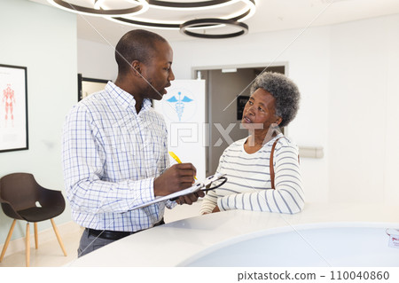 African american male doctor taking notes and talking with senior woman in hospital reception 110040860