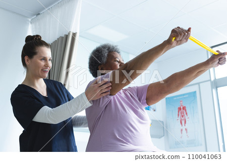 Diverse senior female patient exercising with band and female doctor advising in hospital room 110041063