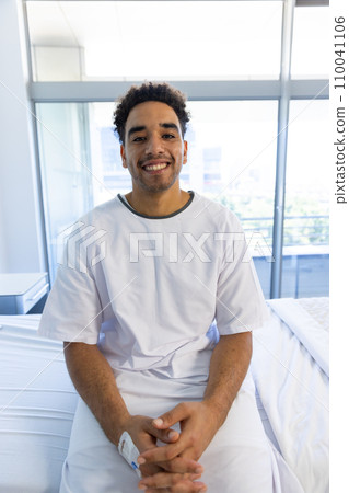 Portrait of happy biracial male patient sitting on bed with iv drip in sunny hospital room 110041106