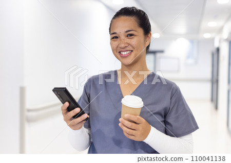 Portrait of happy biracial female doctor using smartphone, holding takeaway coffee in hospital 110041138