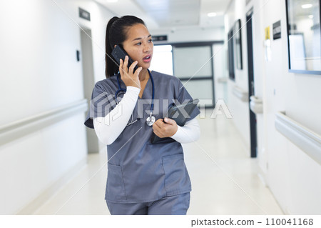 Biracial female doctor wearing scrubs talking on smartphone in hospital corridor Biracial female doctor wearing scrubs talking on smartphone in hospital corridor 110041168