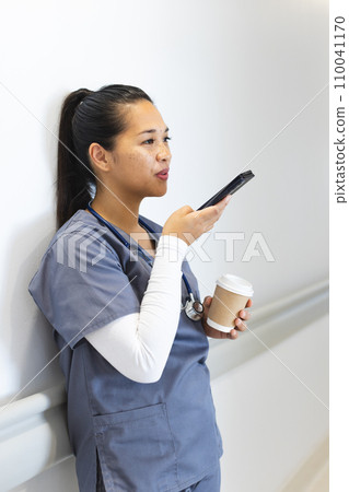 Biracial female doctor talking on smartphone, holding takeaway coffee in hospital corridor Biracial female doctor talking on smartphone, holding takeaway coffee in hospital corridor 110041170