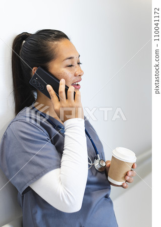 Biracial female doctor talking on smartphone, holding takeaway coffee in hospital corridor 110041172