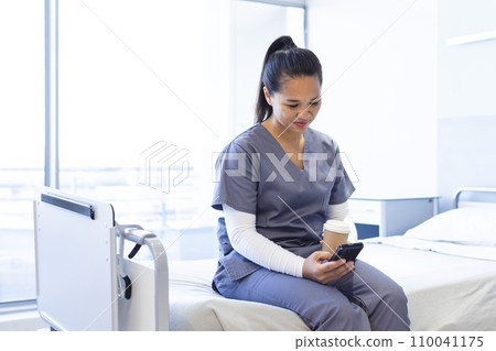 Biracial female doctor using smartphone, holding takeaway coffee on bed in hospital, copy space 110041175