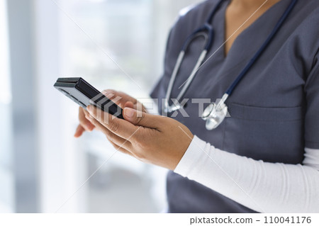 Midsection of biracial female doctor wearing scrubs using smartphon in hospital 110041176