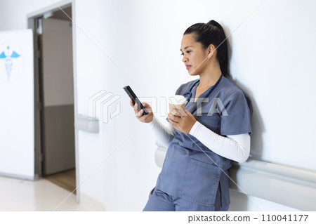 Biracial female doctor wearing scrubs using smartphone, holding takeaway coffee, copy space Biracial female doctor wearing scrubs using smartphone, holding takeaway coffee, copy space 110041177