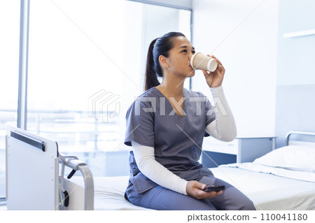 Biracial female doctor using smartphone, drinking takeaway coffee on bed in hospital, copy space Biracial female doctor using smartphone, drinking takeaway coffee on bed in hospital, copy space 110041180
