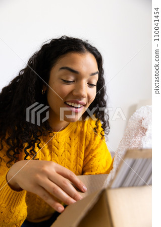 Happy biracial woman in yellow sweater opening package at home Happy biracial woman in yellow sweater opening package at home 110041454