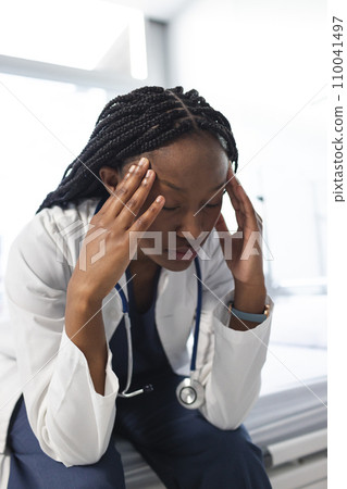 Tired african american female doctor wearing lab coat sitting on bed in hospital room 110041497