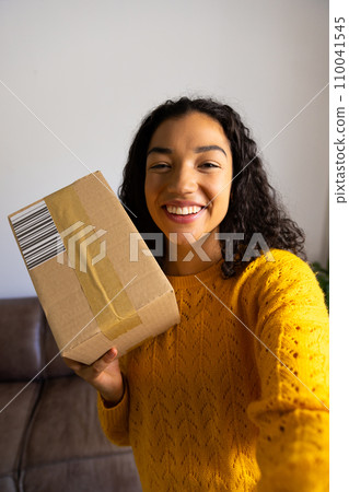 Happy biracial woman in yellow sweater holding package at home 110041545