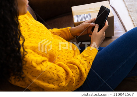 Midsection of biracial woman in yellow sweater with package using smartphone at home Midsection of biracial woman in yellow sweater with package using smartphone at home 110041567