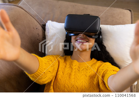 Happy biracial woman lying on sofa and using vr headset at home Happy biracial woman lying on sofa and using vr headset at home 110041568