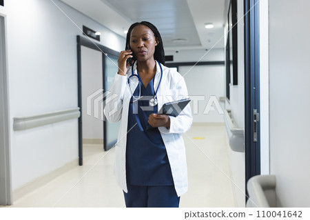 African american female doctor wearing lab coat talking on smartphone in hospital corridor 110041642