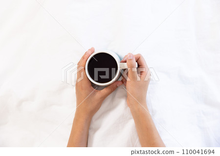 Hands of biracial woman holding cup of coffee sitting in bed at sunny home Hands of biracial woman holding cup of coffee sitting in bed at sunny home 110041694