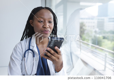 African american female doctor wearing lab coat using smartphone in hospital 110041706