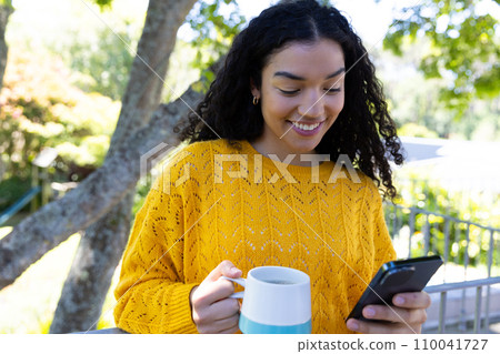 Happy biracial woman in yellow sweater holding cup of coffee and using smartphone on sunny terrace 110041727