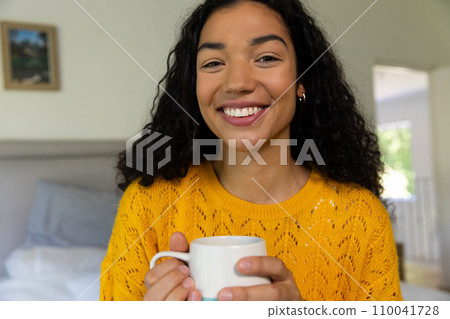 Happy biracial woman in yellow sweater holding cup of coffee at home 110041728