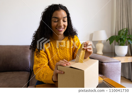 Happy biracial woman in yellow sweater opening package at home 110041730