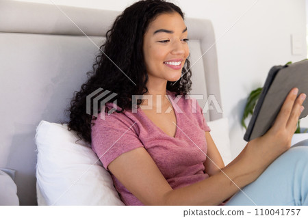 Happy biracial woman using tablet sitting on bed at sunny home 110041757