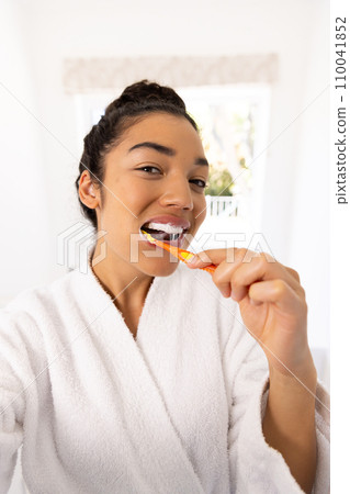 Portrait of biracial woman in bathrobe brushing teeth in sunny bathroom 110041852