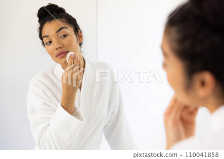 Biracial woman in bathrobe cleansing her face in sunny bathroom Biracial woman in bathrobe cleansing her face in sunny bathroom 110041858
