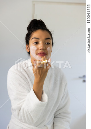 Biracial woman in bathrobe applying face mask using brush in sunny bathroom Biracial woman in bathrobe applying face mask using brush in sunny bathroom 110041863