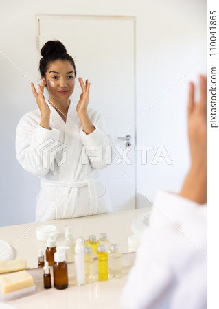 Biracial woman in bathrobe applying cream on her face in sunny bathroom Biracial woman in bathrobe applying cream on her face in sunny bathroom 110041865