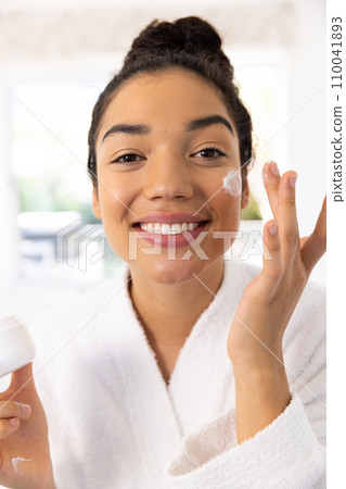 Portrait of happy biracial woman in bathrobe applying cream on her face in sunny bathroom Portrait of happy biracial woman in bathrobe applying cream on her face in sunny bathroom 110041893