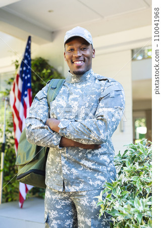 Happy african american male soldier with arms crossed in cap outside home with usa flag, copy space Happy african american male soldier with arms crossed in cap outside home with usa flag, copy space 110041968