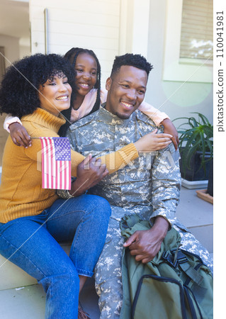 Happy african american male soldier embracing family outside home with usa flag, copy space Happy african american male soldier embracing family outside home with usa flag, copy space 110041981