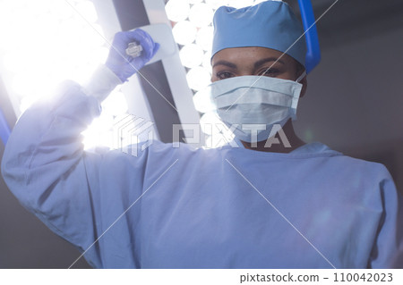 Portrait of biracial female surgeon wearing surgical gown and face mask in operating theatre 110042023