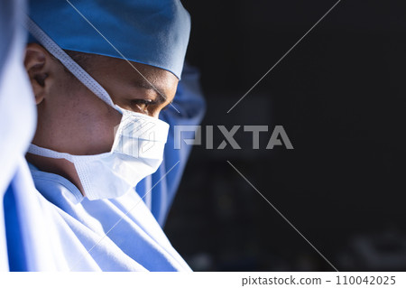 Biracial female surgeon wearing surgical gown and face mask in operating theatre, copy space 110042025