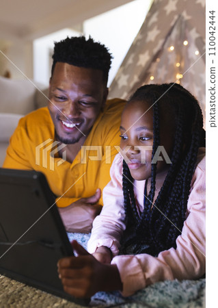 Happy african american father and daughter lying by teepee with tablet at home, copy space 110042444