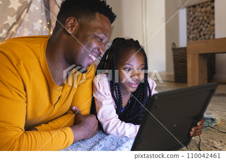 Happy african american father and daughter lying by teepee with computer at home, copy space 110042461