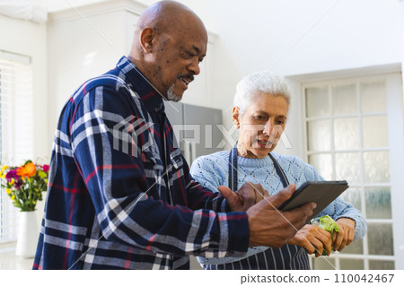 Diverse senior couple preparing healthy meal with vegetables using tablet in kitchen 110042467