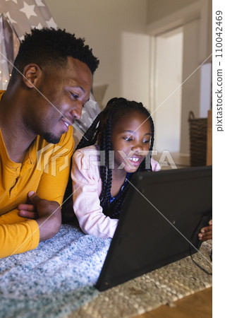 Happy african american father and daughter lying by teepee with tablet at home, copy space 110042469