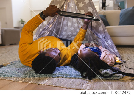 Happy african american father and daughter lying by teepee with tablet at home, copy space 110042472