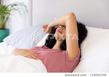 Tired biracial woman holding hand on her head lying in bed at sunny home Tired biracial woman holding hand on her head lying in bed at sunny home 110042546