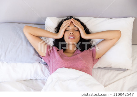 Tired biracial woman holding hands on her head lying in bed at sunny home 110042547