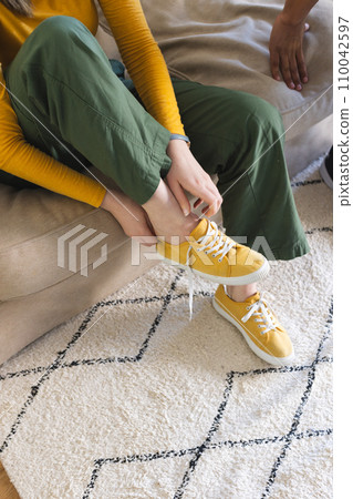 Caucasian woman sitting on couch and putting yellow shoes on in living room at home, copy space 110042597