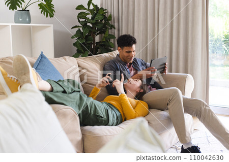 Happy diverse couple lying on couch, using tablet and smartphone in living room at home, copy space 110042630
