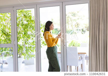 Thoughtful caucasian woman standing by window and holding tea cup in living room at home, copy space 110042631